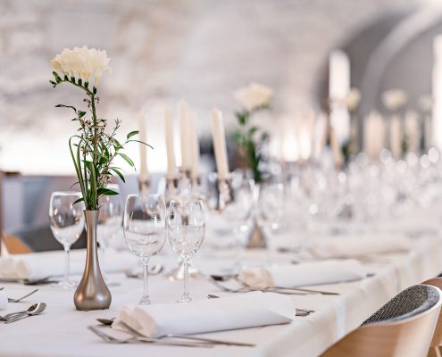 Ein vollständig eingedeckter langer Tisch mit weißer Tischdecke, silbernen Fasen mit weißen Blumen darin und Kerzenständer mit weißen Kerzen.