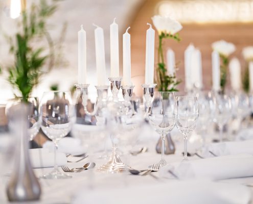 Ein vollständig eingedeckter langer Tisch mit weißer Tischdecke, silbernen Fasen mit weißen Blumen darin und Kerzenständer mit weißen Kerzen, welche im Fokus stehen.